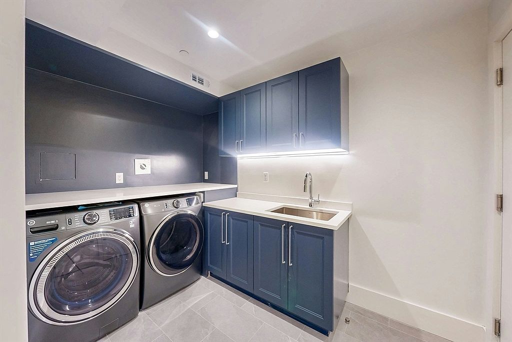 This is a well-organized laundry room featuring a front-loading washer and dryer set, blue cabinetry with silver hardware, and a white countertop with a stainless steel sink. The room is painted in neutral tones, with a dark blue accent wall above the washer and dryer, and the flooring is a light gray tile. The overall impression is clean, modern, and functional.