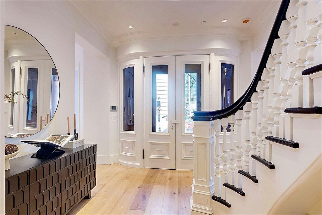 This is an interior shot of a grand entryway featuring a staircase with white balusters and a dark wood handrail. A dark wood console table with decorative items sits to the left, complemented by a large round mirror. The entrance doors are white with glass panels, allowing natural light to flood the space, highlighting the light wood flooring.