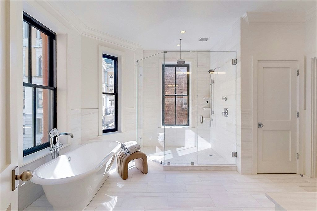 This is a bright and luxurious primary bathroom featuring a freestanding bathtub positioned beneath a large window, a glass-enclosed shower with modern fixtures, and white marble tiling throughout. The room is well-lit with natural light and recessed lighting, creating a clean and spa-like atmosphere. The black window frames add a modern contrast to the otherwise white color palette.