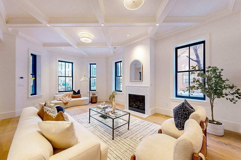 This is an interior shot of a living room featuring a white sofa with decorative pillows, two armchairs, and a glass coffee table on a patterned rug. The room has a fireplace with a mirror above it, and several windows with black frames that let in natural light. The overall style is modern and elegant, with a focus on clean lines and neutral colors.