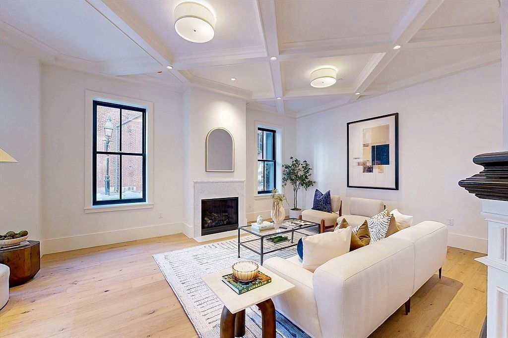 This is an interior shot of a living room featuring a coffered ceiling with recessed lighting and two modern drum light fixtures. The room is furnished with a light-colored sofa, armchairs, a glass-topped coffee table, and a side table. A fireplace with a marble surround and a large framed artwork add to the room's elegance, while the hardwood floors and area rug provide warmth and texture.