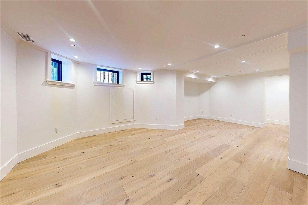 This is a spacious, finished basement featuring light hardwood flooring and white walls. Three small windows provide natural light, and recessed lighting illuminates the room. The space appears clean and modern, offering a versatile area for various uses.