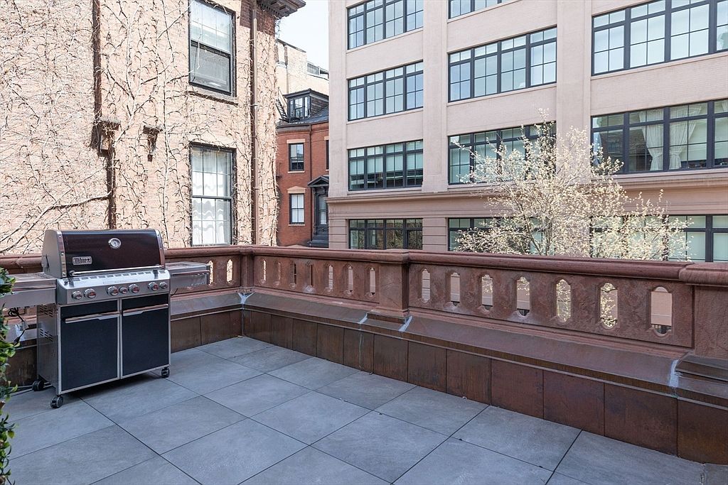 This image showcases a spacious outdoor patio or balcony area, featuring a modern grill, tiled flooring, and a decorative stone railing. The backdrop includes a mix of architectural styles, with buildings featuring brick and stone facades, creating an urban yet inviting atmosphere. The perspective is from a slightly elevated angle, providing a comprehensive view of the outdoor space and its surroundings.