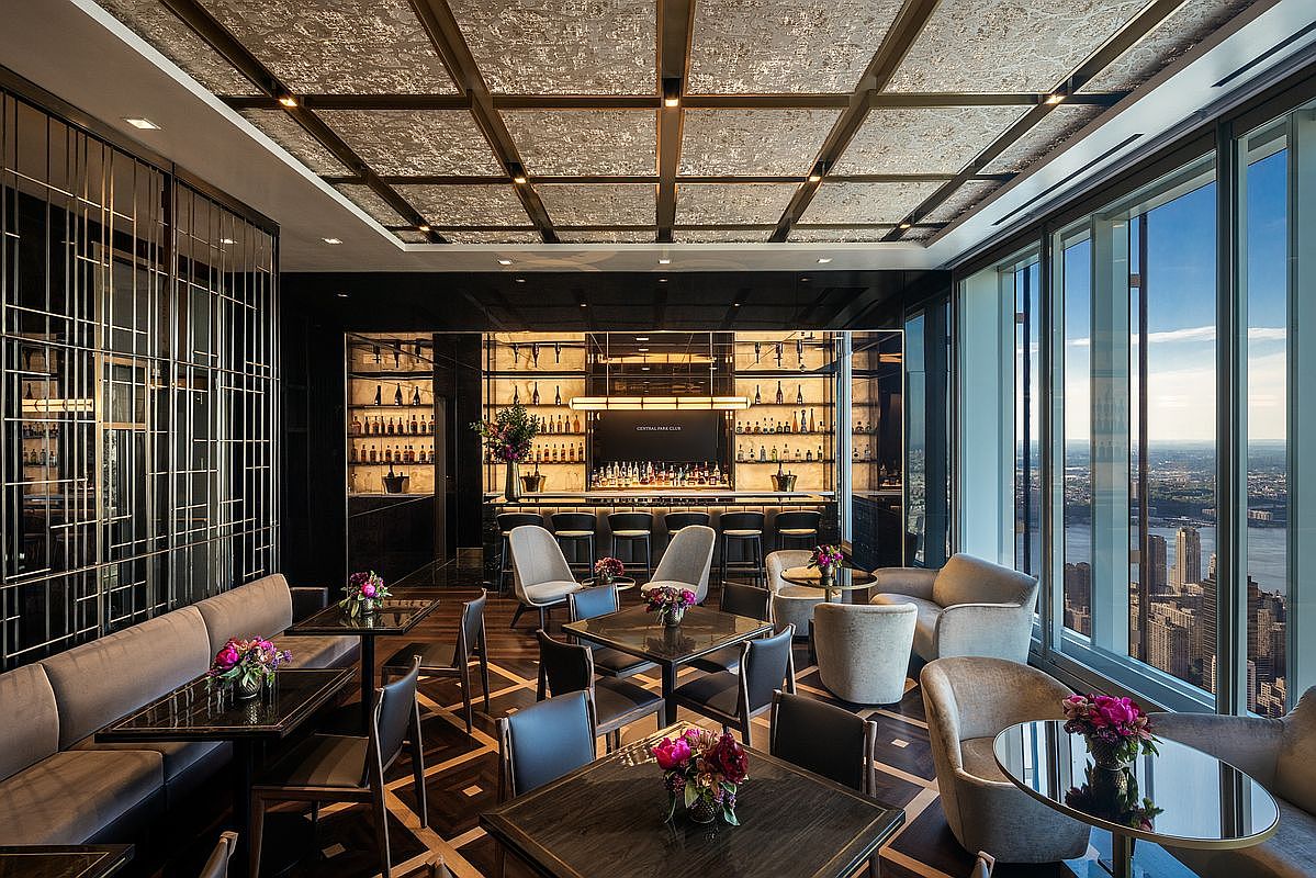 This sophisticated high-rise lounge features a luxurious, modern aesthetic with a coffered ceiling, dark wood accents, and plush seating arrangements. A sleek bar area serves as the focal point, while floor-to-ceiling windows provide a breathtaking, panoramic view of the city skyline. The space is elegantly decorated with floral arrangements, creating an upscale atmosphere perfect for social gatherings.