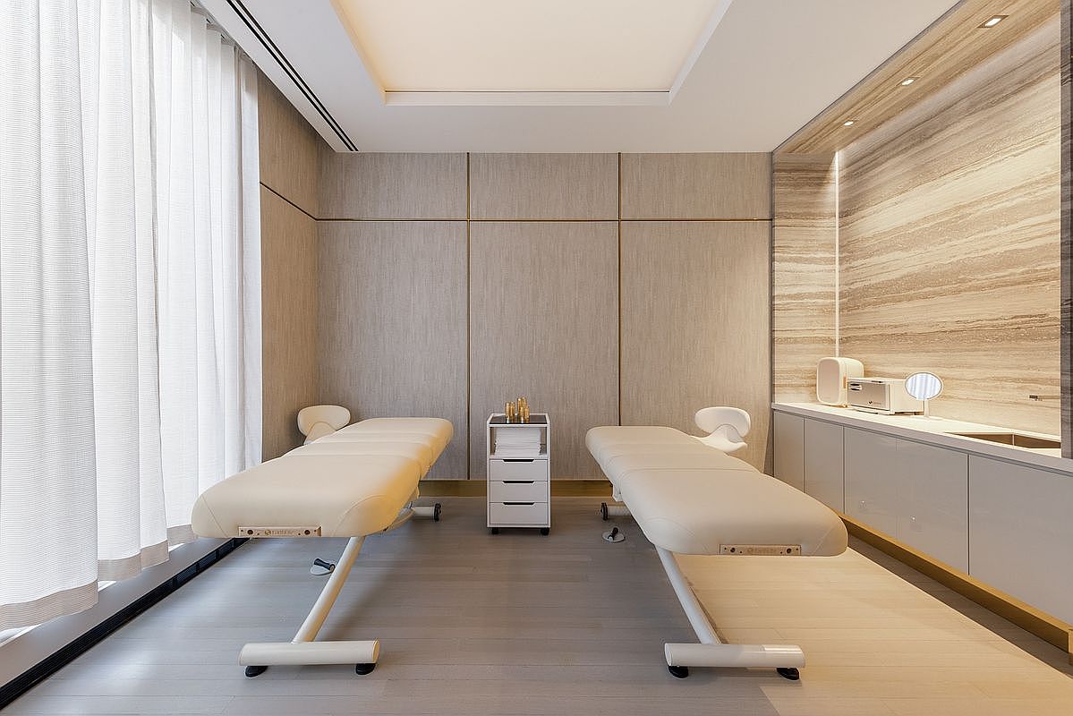 This serene and minimalist spa treatment room features two professional massage tables positioned symmetrically on a light-toned wood floor. The space is accented by textured wall paneling, a sleek built-in vanity with marble-like backsplash, and soft, diffused lighting that creates a calming atmosphere. A sheer curtain on the left allows for gentle natural light, enhancing the clean, high-end aesthetic of this wellness facility.
