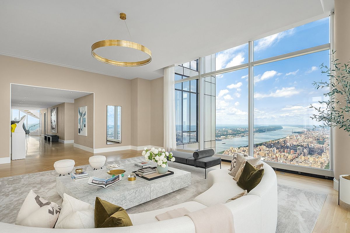 This sophisticated, high-rise living room features a curved white sofa, a large marble coffee table, and a striking gold circular chandelier. Floor-to-ceiling windows provide a breathtaking, panoramic view of the city skyline and river, creating an airy and luxurious atmosphere. The neutral color palette and minimalist decor emphasize the expansive space and the dramatic urban backdrop.