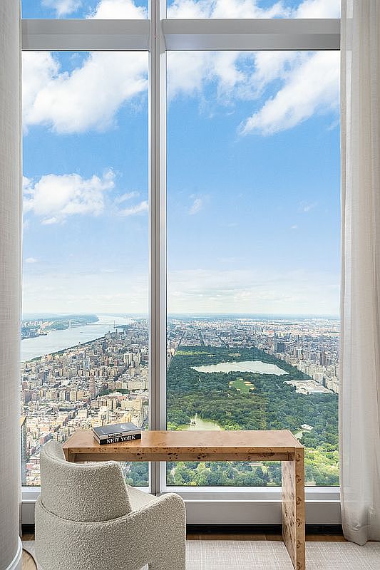 This sophisticated home office space features a minimalist wooden desk positioned directly against a floor-to-ceiling window, offering a breathtaking, unobstructed aerial view of Central Park and the New York City skyline. A plush, textured cream-colored armchair provides a comfortable seating option, while sheer white curtains frame the expansive glass panels. The overall aesthetic is one of luxury and serenity, emphasizing the dramatic urban panorama as the primary focal point of the room.
