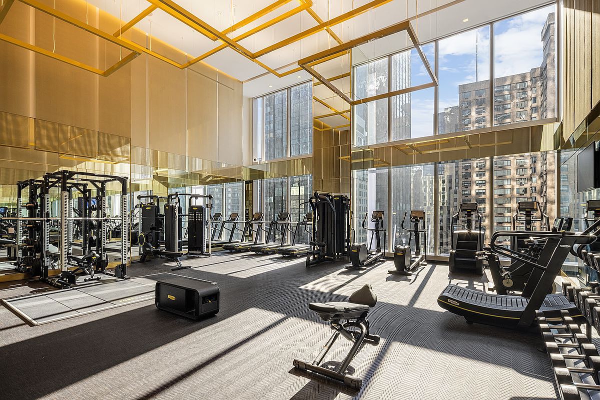 This high-end, modern fitness center features floor-to-ceiling windows that offer expansive city views and bathe the space in natural light. The room is equipped with professional-grade Technogym equipment, including a power rack, treadmills, and elliptical machines, all arranged on a sleek, dark herringbone-patterned floor. Gold-toned accents and mirrored walls enhance the luxurious, sophisticated atmosphere of this premium residential amenity.