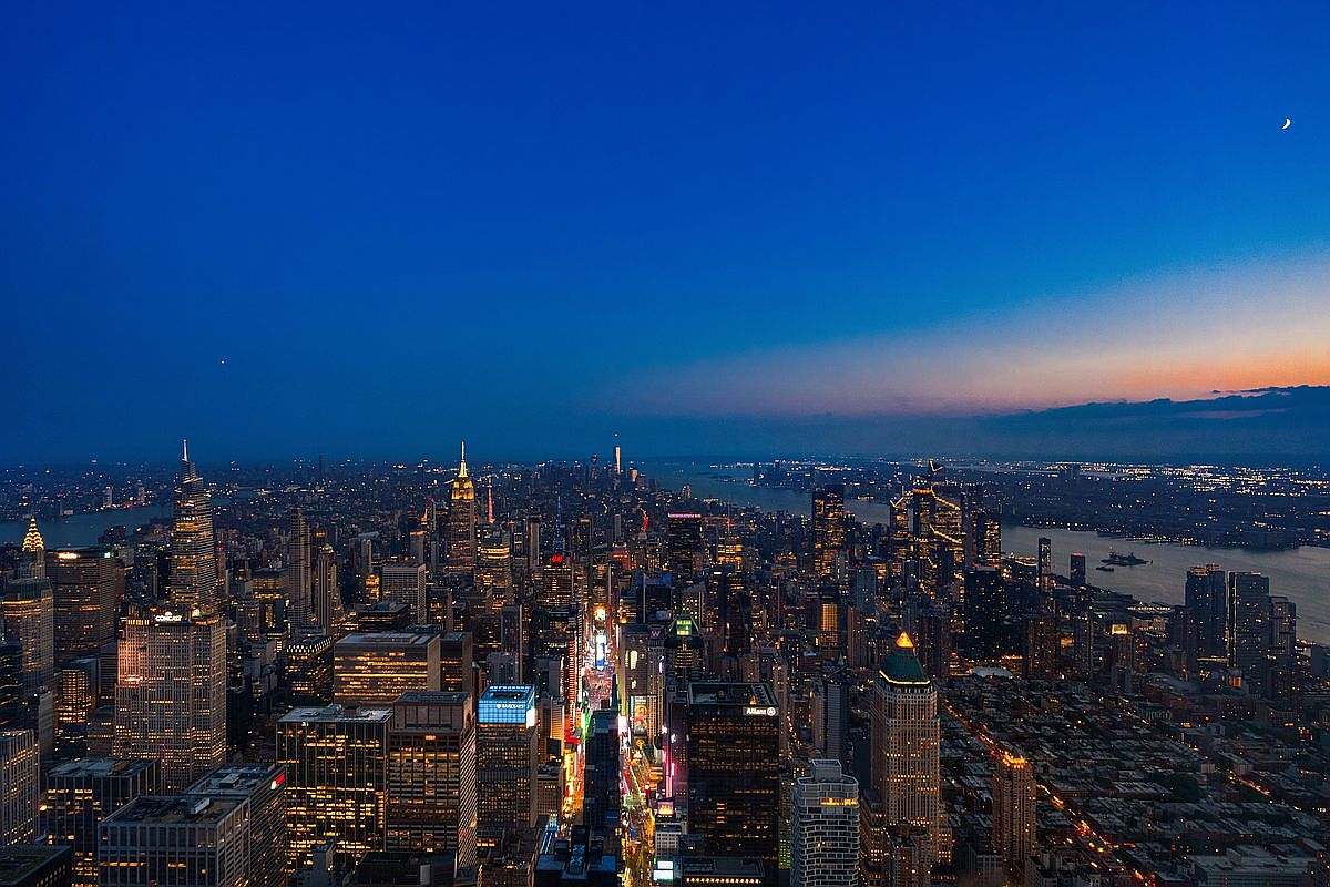 This breathtaking high-altitude aerial view captures the iconic New York City skyline at twilight, showcasing a dense urban landscape illuminated by thousands of glowing windows and streetlights. The perspective highlights major architectural landmarks, including the Empire State Building and the vibrant grid of Times Square, set against a deep blue dusk sky with a crescent moon. The composition offers a cinematic, expansive look at the city's scale and density, emphasizing its status as a premier global metropolis.