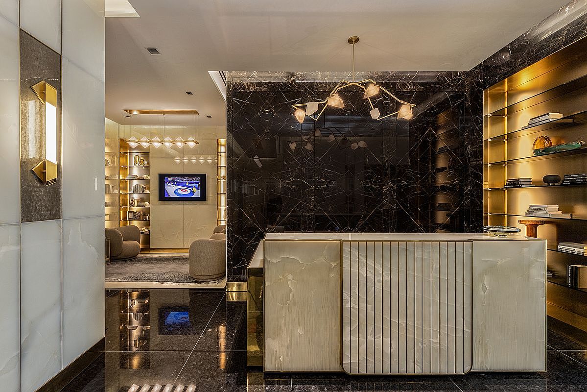 This sophisticated interior space features a striking reception area defined by a polished light-stone desk with vertical gold accents, set against a dramatic black marble wall. The perspective looks through a grand, open-concept hallway into a secondary lounge area, showcasing a cohesive design language of luxury materials, warm gold lighting, and high-end finishes. The overall impression is one of opulent, modern elegance suitable for a high-end commercial lobby or a luxury residential foyer.