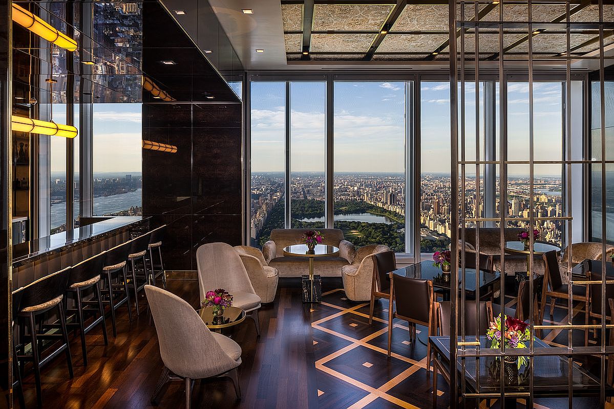 This sophisticated high-rise lounge offers a luxurious interior space with floor-to-ceiling windows providing a breathtaking panoramic view of Central Park and the New York City skyline. The room is elegantly furnished with plush velvet armchairs, dark wood flooring with geometric inlay, and a sleek bar area, creating an atmosphere of refined exclusivity. The cinematic perspective captures the seamless blend of opulent indoor design and the iconic urban landscape beyond.