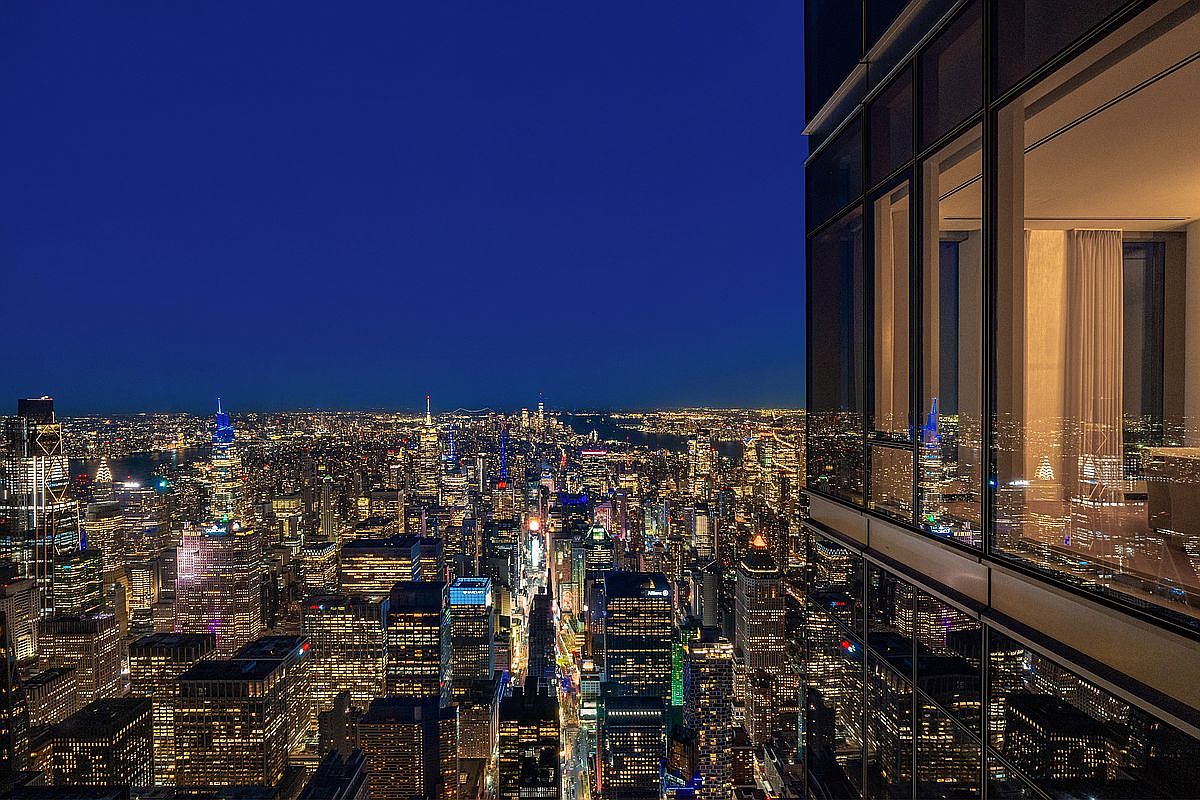 This striking aerial view captures a high-rise perspective of the New York City skyline at twilight, showcasing a dense urban landscape illuminated by thousands of city lights. In the foreground, the sleek, modern glass facade of the building provides a reflective frame, offering a glimpse into a luxurious interior space. The composition emphasizes the dramatic contrast between the vast, glowing cityscape and the sophisticated, quiet interior environment.