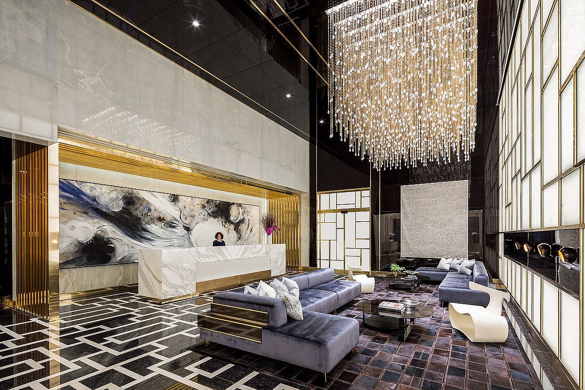 This luxurious lobby features a grand, double-height ceiling adorned with a massive, cascading crystal chandelier that serves as the room's focal point. The space is elegantly furnished with plush, velvet-upholstered sofas, a sleek marble reception desk, and sophisticated geometric flooring, all accented by warm gold trim and high-end finishes. The overall aesthetic is one of opulent, contemporary refinement, designed to impress residents and guests alike.
