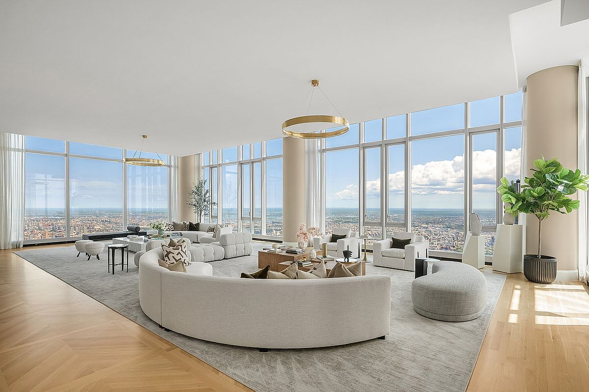 This expansive, high-floor living room features a sophisticated open-concept layout with floor-to-ceiling windows that offer panoramic city views. The space is anchored by a large, curved cream-colored sofa and a secondary modular seating arrangement, all set atop a plush, neutral-toned area rug. The interior design is modern and minimalist, highlighted by elegant gold circular chandeliers and light hardwood flooring, creating an atmosphere of refined luxury.