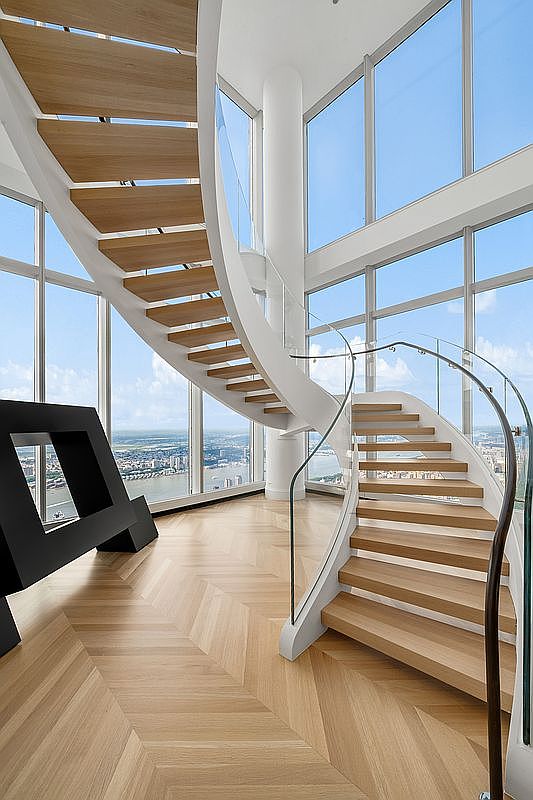 This stunning interior space features a dramatic, sculptural spiral staircase with light wood treads and a sleek white frame, serving as the centerpiece of the room. Floor-to-ceiling glass windows provide an expansive, panoramic view of the city skyline and river below, bathing the room in natural light. The herringbone-patterned hardwood flooring and minimalist aesthetic create a sophisticated, high-end atmosphere typical of a luxury penthouse.
