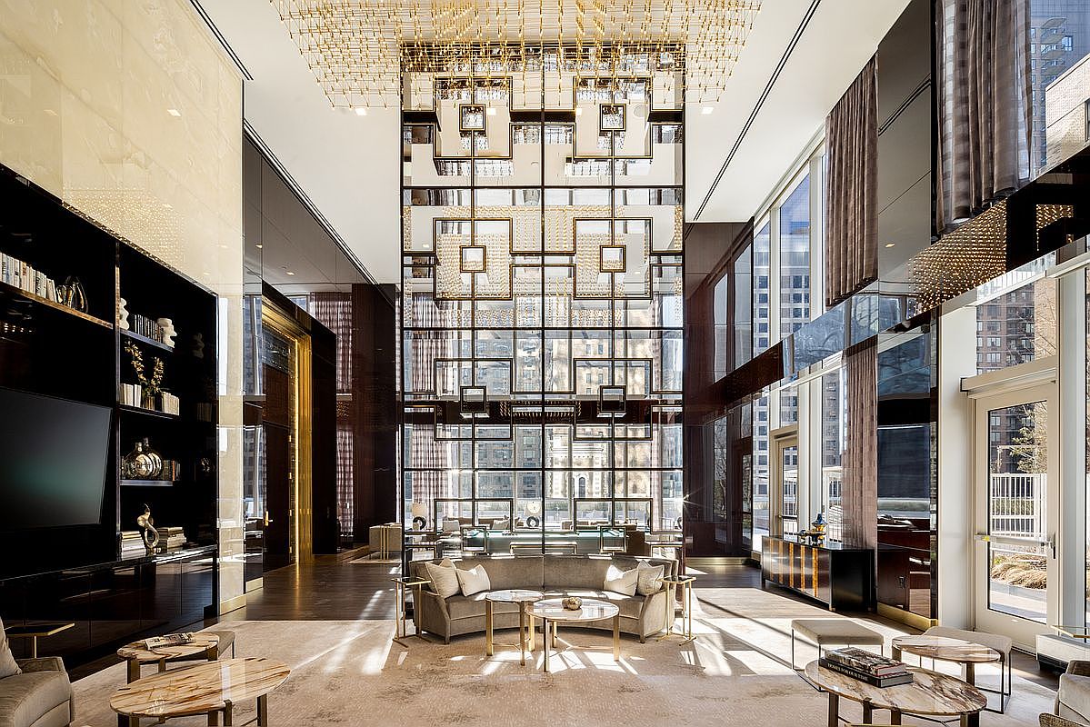 This grand, double-height lobby or lounge area exudes luxury with its soaring ceilings, intricate geometric metal screen, and a massive, ornate gold-toned chandelier. The space is furnished with plush, contemporary seating, marble-topped coffee tables, and a sleek built-in media wall, creating an atmosphere of sophisticated elegance. The perspective is a wide, symmetrical shot that emphasizes the architectural scale and high-end finishes of this upscale residential amenity space.