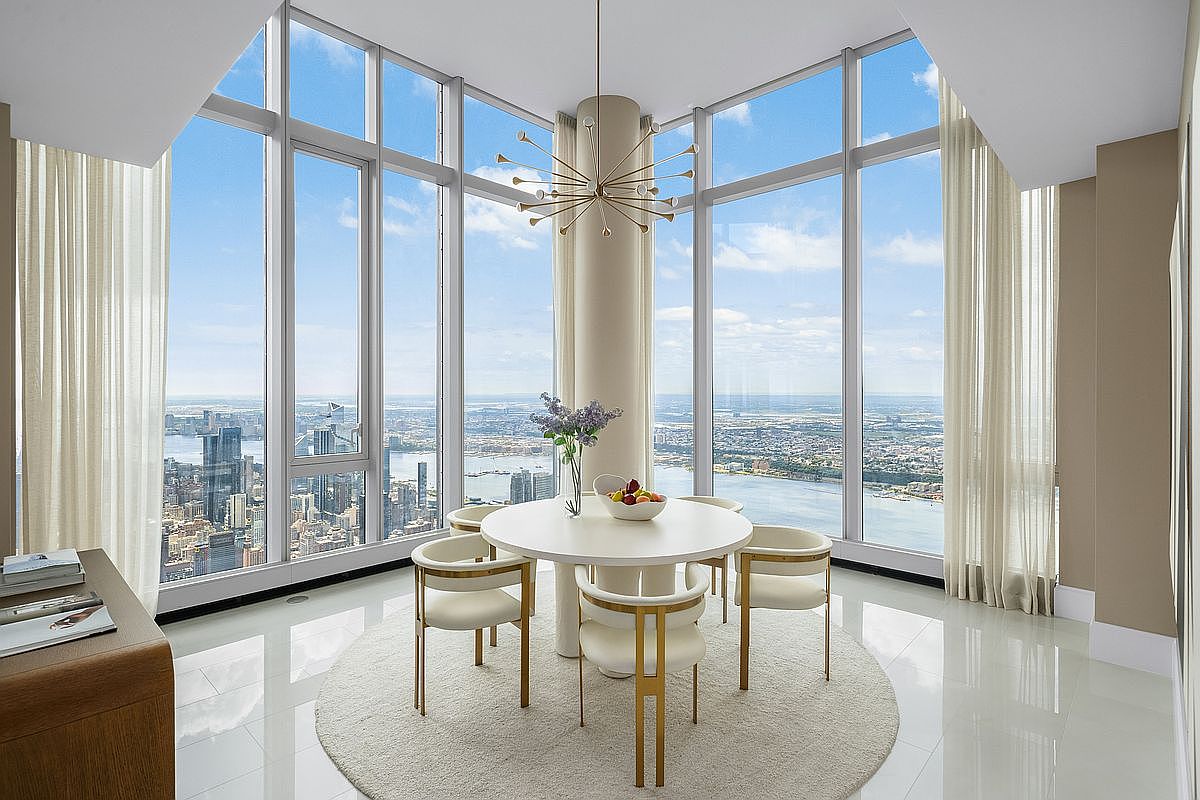 This sophisticated dining area features a minimalist white circular table surrounded by four elegant, gold-accented chairs, all set atop a plush, neutral-toned area rug. The space is defined by floor-to-ceiling windows that offer a breathtaking, panoramic view of the city skyline and river, while a striking sputnik-style chandelier hangs from the ceiling as a modern focal point. The room exudes a luxurious, airy atmosphere, perfectly blending contemporary design with an unparalleled urban backdrop.