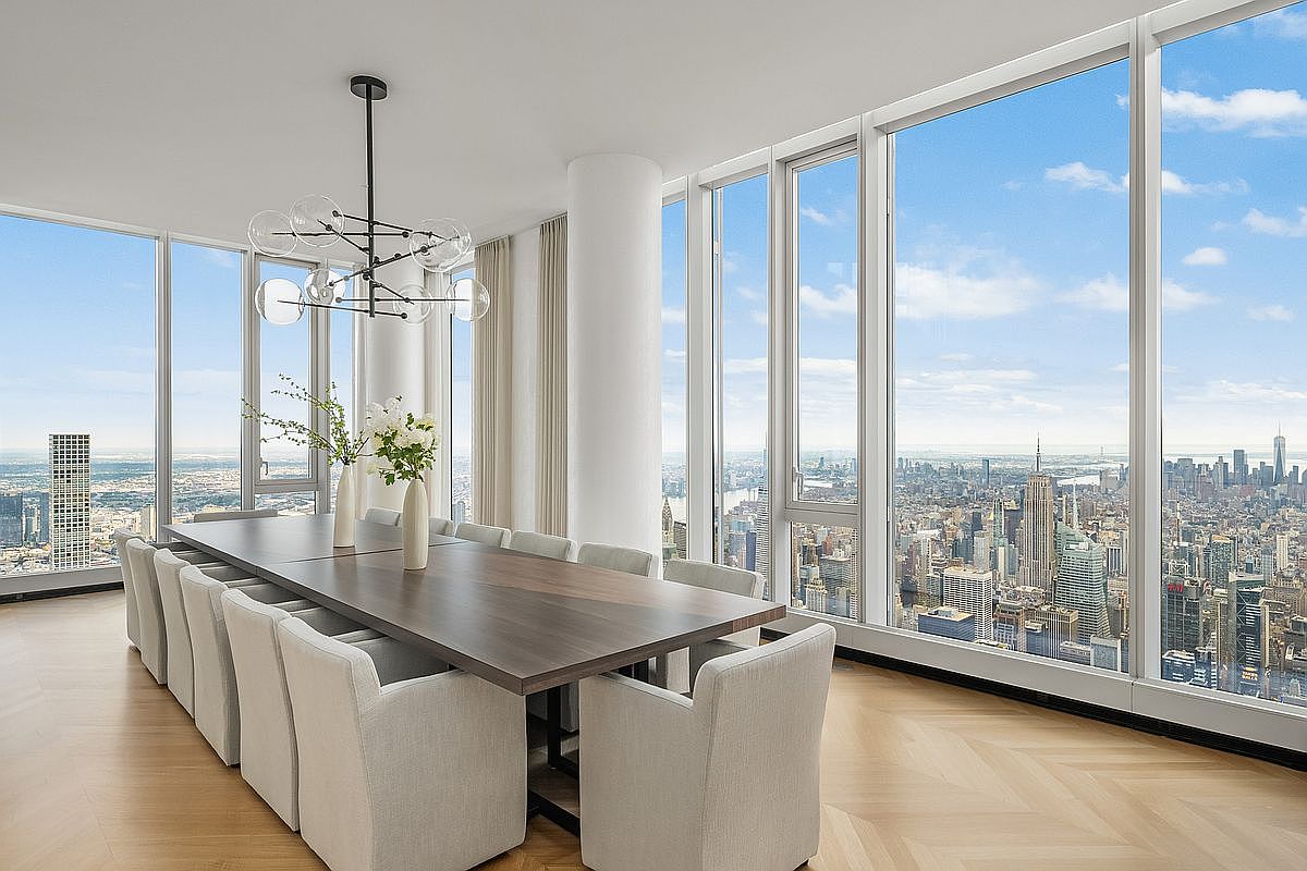 This sophisticated dining room features a long, dark wood table surrounded by light-colored upholstered chairs, set against a backdrop of floor-to-ceiling windows that offer a breathtaking panoramic view of the New York City skyline. A modern, multi-globe chandelier hangs above the table, adding a touch of contemporary elegance to the bright, airy space with its light hardwood floors. The room exudes a sense of luxury and high-rise living, perfectly framing the iconic urban landscape.