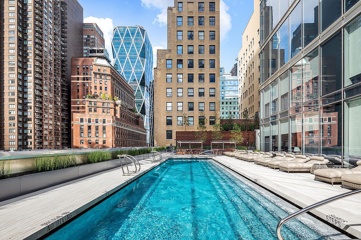 This stunning rooftop lap pool offers a luxurious urban oasis, framed by a sleek wooden deck and rows of comfortable lounge chairs. The perspective captures the pool stretching toward a backdrop of iconic New York City architecture, including the distinctive Hearst Tower. The scene exudes a sophisticated, high-end metropolitan lifestyle, perfect for relaxation amidst the bustling city skyline.