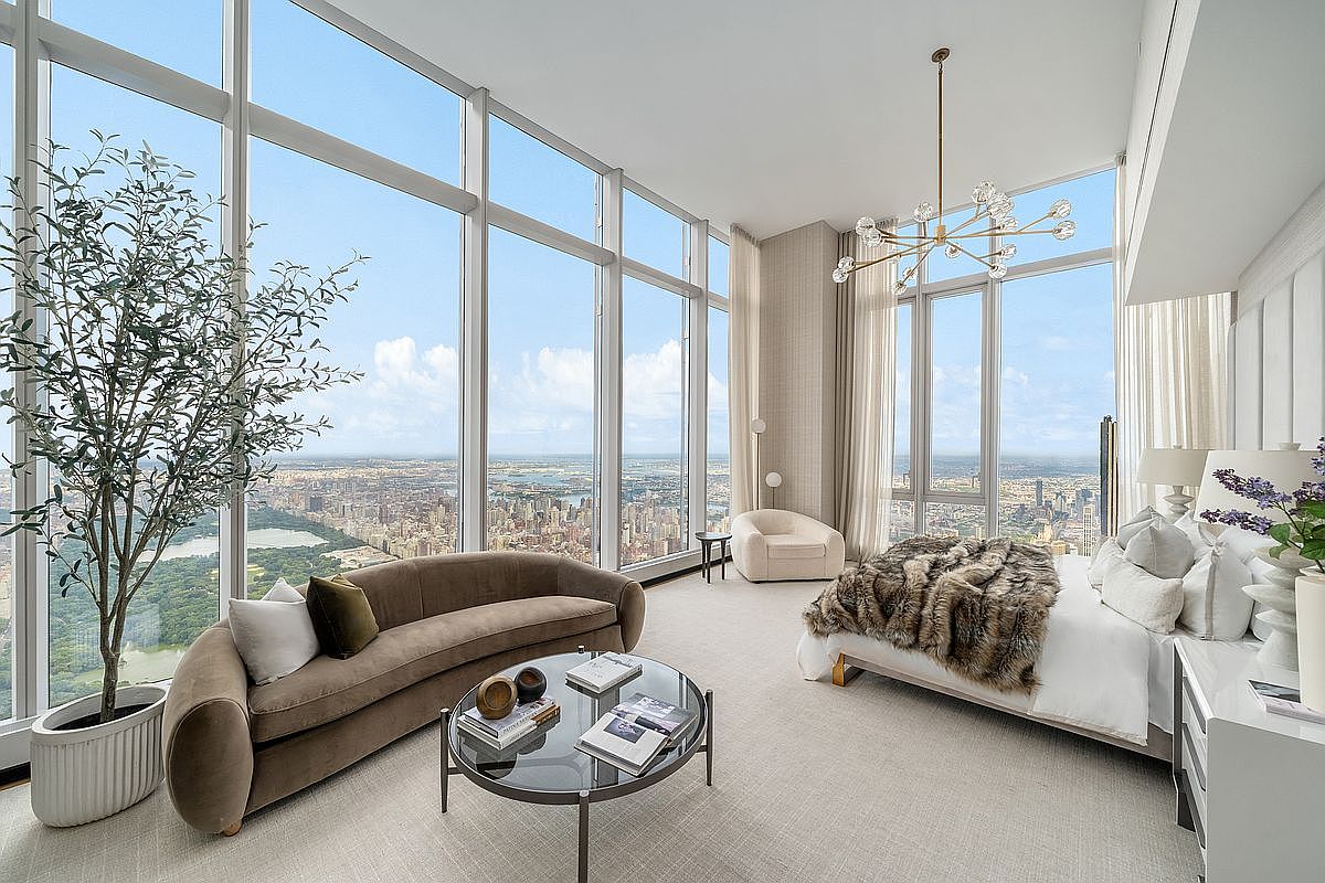 This luxurious primary bedroom features floor-to-ceiling windows that offer a breathtaking, panoramic view of the city skyline and Central Park. The room is elegantly furnished with a plush, curved sofa, a modern glass coffee table, and a bed adorned with a textured faux-fur throw, all set against a neutral, sophisticated color palette. The high ceilings and minimalist decor emphasize the expansive, airy feel of this high-rise residence.