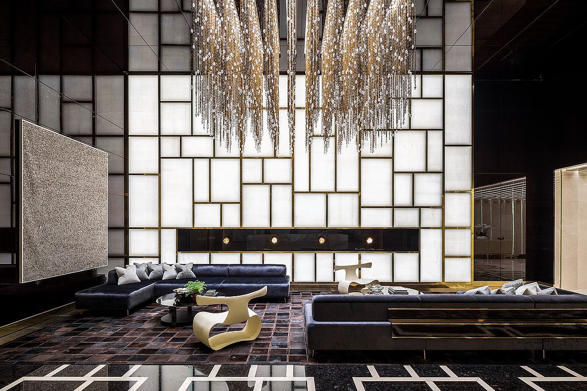 This ultra-luxurious, high-ceilinged living room features a dramatic, floor-to-ceiling backlit geometric wall panel and a cascading crystal chandelier that serves as a stunning focal point. The space is furnished with plush, dark velvet sectional sofas, sculptural modern chairs, and a rich, textured area rug, all set against a backdrop of dark, reflective surfaces. The overall aesthetic is one of sophisticated, high-end contemporary design, perfect for an upscale urban residence.