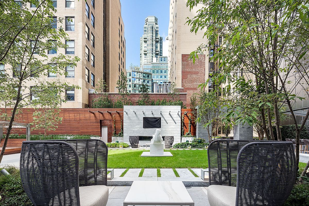 This sophisticated urban garden features a manicured lawn, a central stone sculpture, and a modern fireplace wall, creating a serene oasis amidst high-rise buildings. The foreground is framed by two elegant, dark-woven outdoor chairs and a sleek table, inviting relaxation in this private outdoor space. The composition offers a cinematic perspective, looking past the seating area toward the lush greenery and the striking architectural contrast between the garden and the surrounding city skyline.