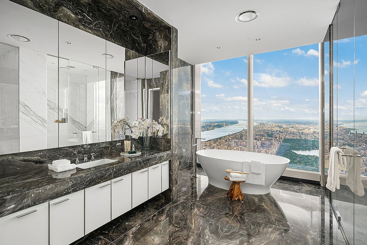 This luxurious primary bathroom features high-end dark marble surfaces, a sleek white vanity with integrated sinks, and a freestanding soaking tub positioned against floor-to-ceiling windows. The panoramic view of the city skyline and river creates a dramatic, sophisticated atmosphere. The space is defined by its modern, minimalist aesthetic and premium materials, offering a serene and opulent retreat.