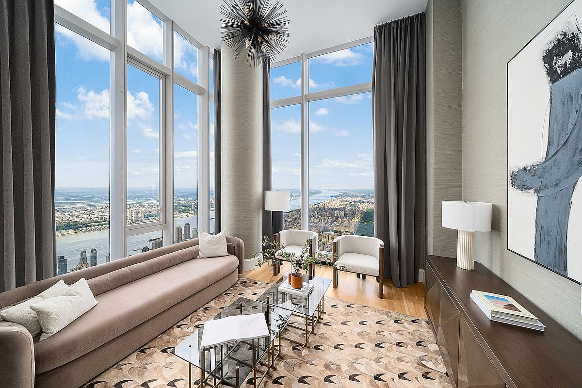 This sophisticated high-rise living room features floor-to-ceiling windows that offer a breathtaking panoramic view of the city skyline and river. The space is elegantly furnished with a long, plush sofa, two modern armchairs, and a unique geometric area rug, all anchored by a striking starburst chandelier. The neutral color palette and clean lines create a luxurious, airy atmosphere perfect for high-end urban living.