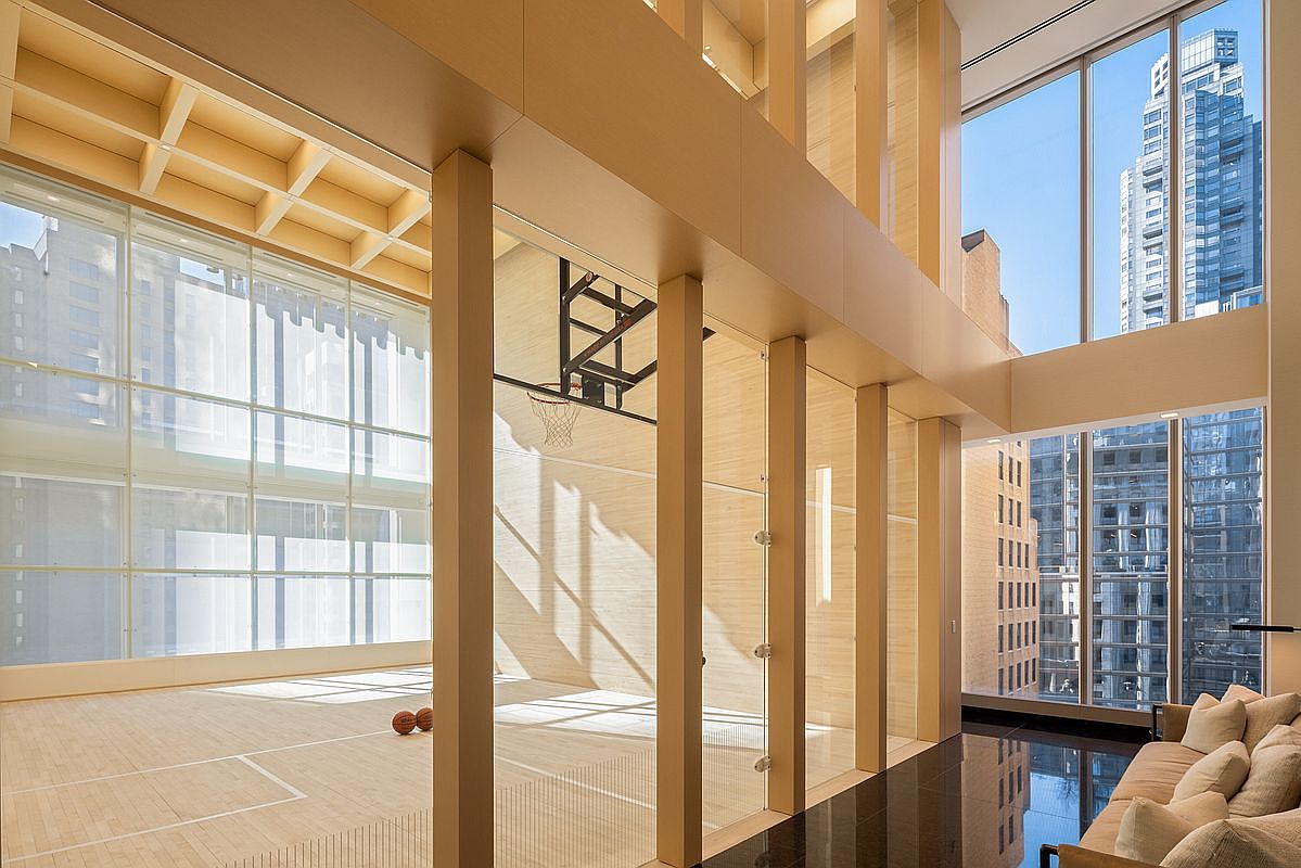 This sophisticated indoor basketball court features a light wood floor and high ceilings, enclosed by floor-to-ceiling glass walls that provide a seamless view into an adjacent lounge area. The space is bathed in natural light from expansive windows, creating an airy and luxurious atmosphere. The minimalist design and high-end finishes make this a premier recreational amenity within a modern residence.