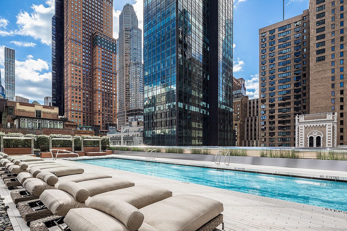 This high-end urban rooftop pool area offers a luxurious retreat amidst a dense cityscape of towering skyscrapers. The scene features a sleek, rectangular swimming pool lined with rows of comfortable, neutral-toned lounge chairs on a clean, light-colored deck. The perspective is a low-angle shot looking across the pool, emphasizing the contrast between the tranquil water and the dramatic, modern architecture surrounding the space.