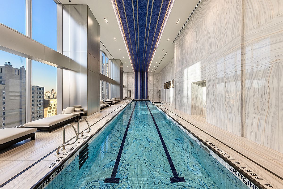 This luxurious indoor lap pool features a stunning mosaic-tiled floor and a dramatic, high-ceilinged design with a striking blue and gold decorative ceiling element. Floor-to-ceiling windows provide expansive city views, while sleek lounge chairs line the side, creating an atmosphere of high-end sophistication and relaxation. The space is finished with premium marble-like wall cladding, emphasizing its status as a premier residential amenity.