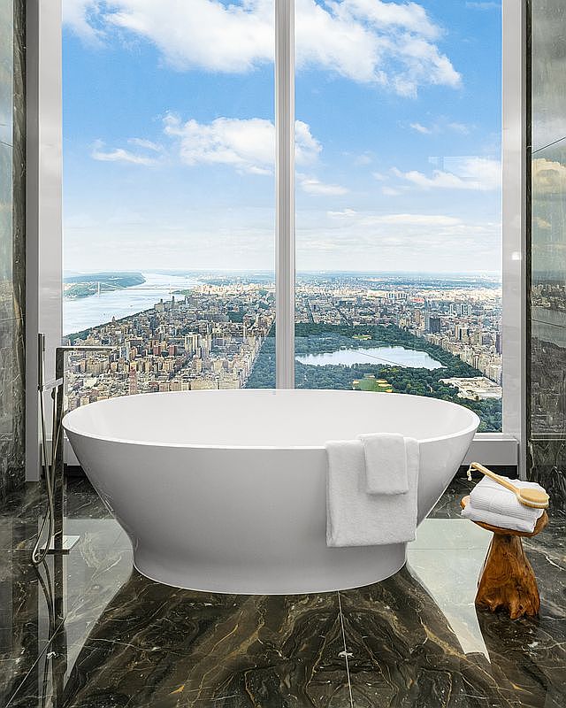This luxurious primary bathroom features a sleek, white freestanding soaking tub positioned perfectly in front of floor-to-ceiling windows that offer a breathtaking, panoramic view of the New York City skyline and Central Park. The room is finished with dramatic, dark veined marble flooring that reflects the natural light, while a rustic wooden stool with folded towels and a bath brush adds a touch of organic warmth to the sophisticated space. The overall impression is one of high-end, modern elegance and unparalleled urban serenity.