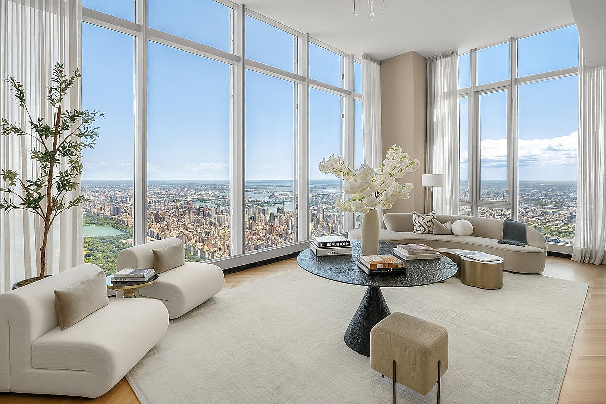 This sophisticated, high-rise living room features floor-to-ceiling windows that offer a breathtaking, panoramic view of the city skyline and Central Park. The space is furnished with elegant, curved cream-colored sofas, a textured circular coffee table, and a minimalist aesthetic that emphasizes luxury and openness. The bright, airy atmosphere is enhanced by neutral tones and a large, soft area rug, creating a serene and upscale urban retreat.