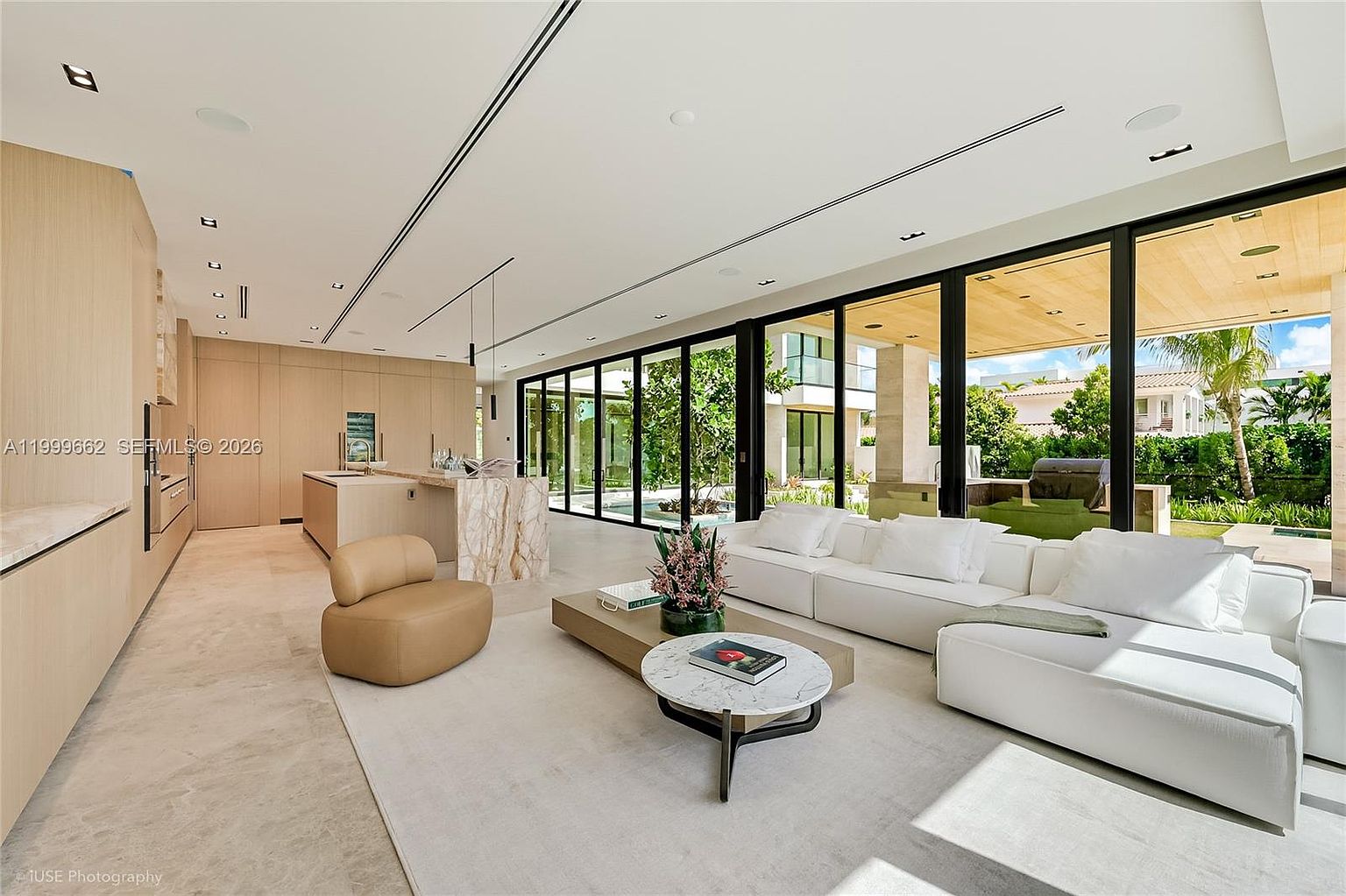 This expansive, open-concept living area features a minimalist aesthetic with light wood cabinetry and neutral-toned furnishings. A large white sectional sofa and a circular marble-topped coffee table anchor the space, while floor-to-ceiling glass walls provide a seamless transition to the outdoor patio and pool area. The room is bathed in natural light, highlighting the clean lines and sophisticated, modern design.
