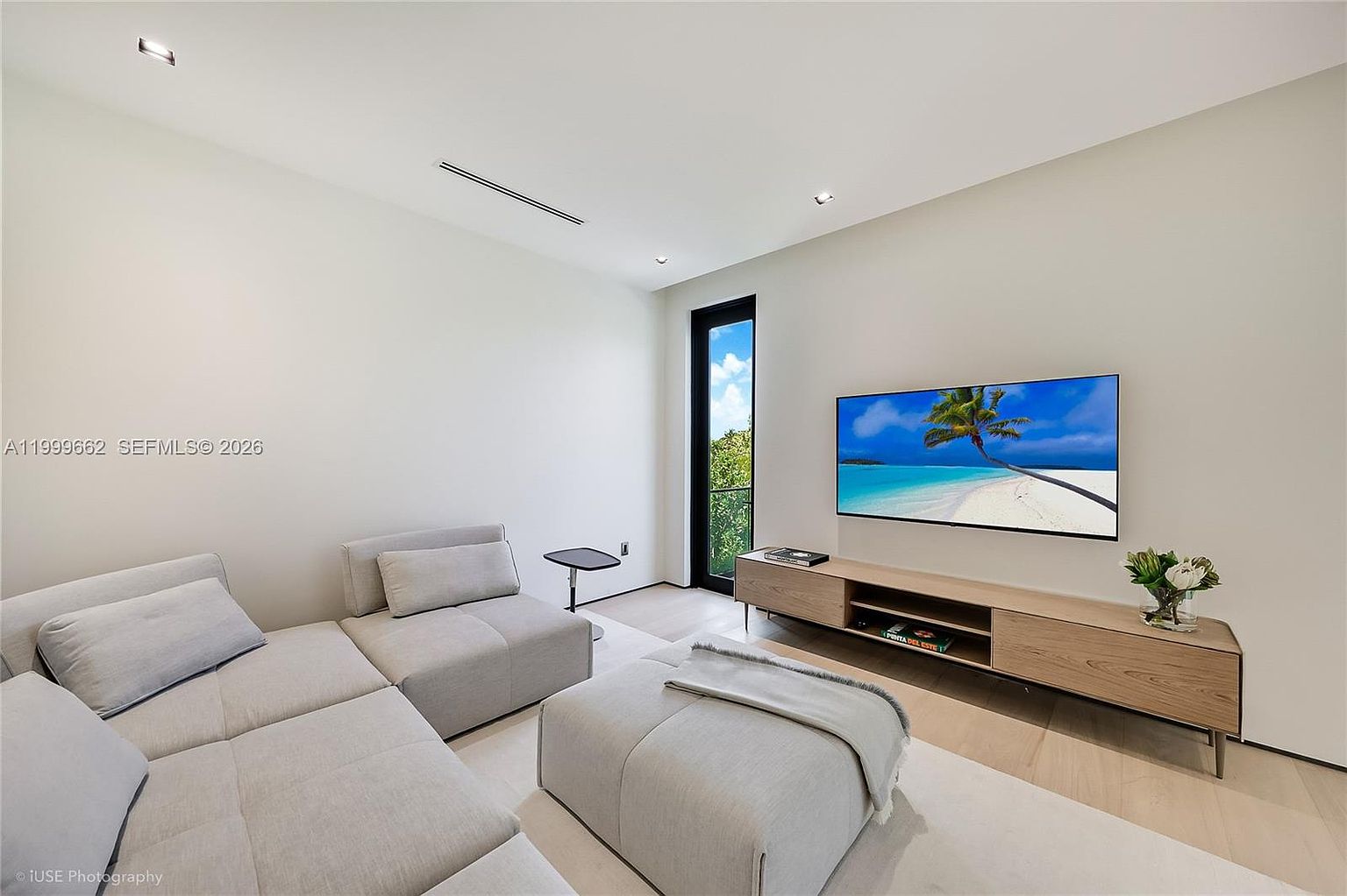 This modern living room features a minimalist aesthetic with light-toned walls and light wood flooring. The space is furnished with a large, modular light gray sectional sofa and a matching ottoman, centered around a sleek, low-profile light wood media console with a wall-mounted television. A tall, narrow window provides natural light and a view of the outdoors, contributing to the bright and airy atmosphere of the room.