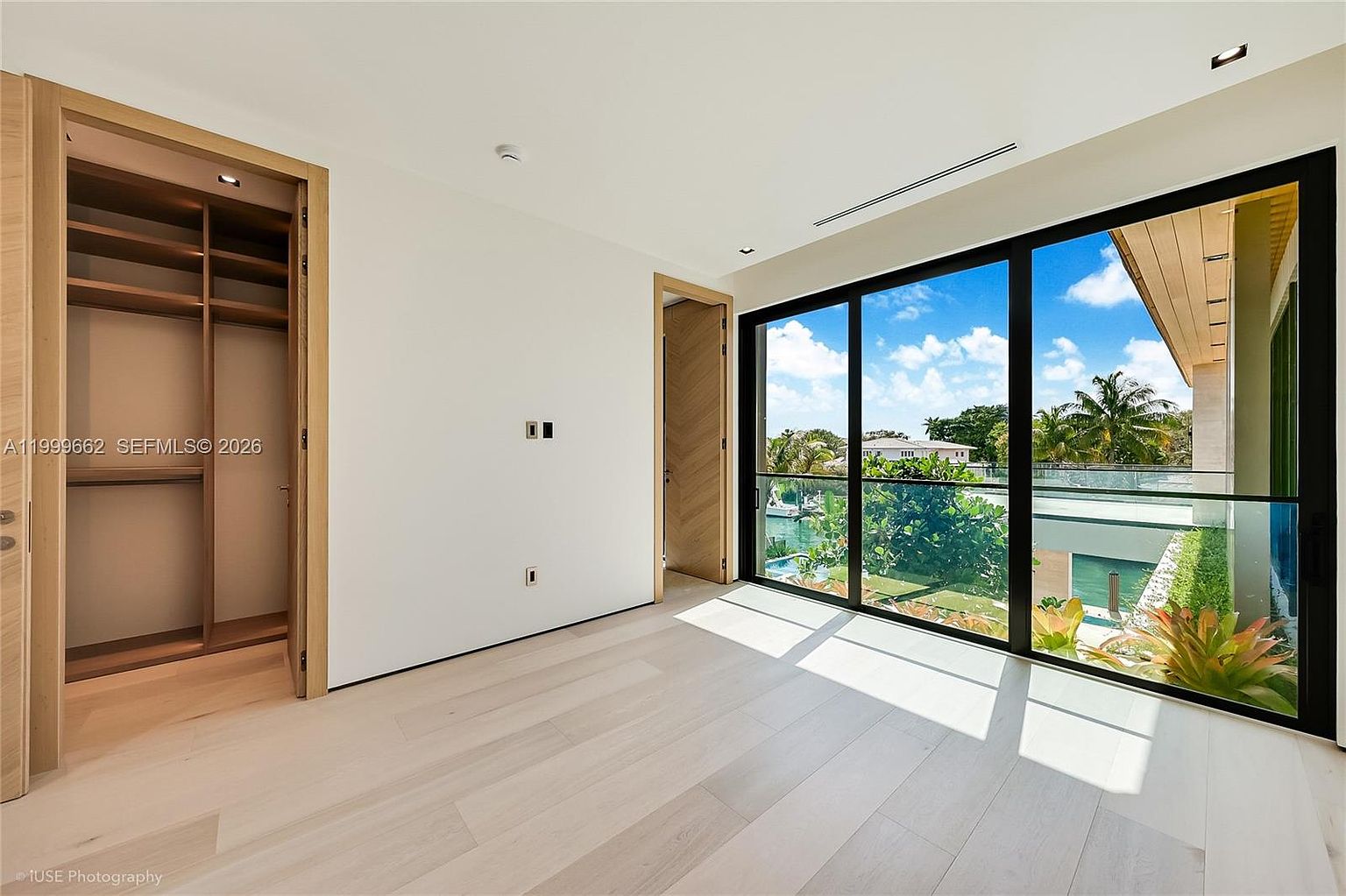 This bright, minimalist bedroom features light-toned wood flooring and crisp white walls, creating an airy and modern atmosphere. A large walk-in closet with built-in shelving is accessible through an open doorway, while expansive glass doors provide a seamless transition to an outdoor balcony with views of the surrounding greenery and water. The room is bathed in natural light, emphasizing its clean lines and contemporary design.