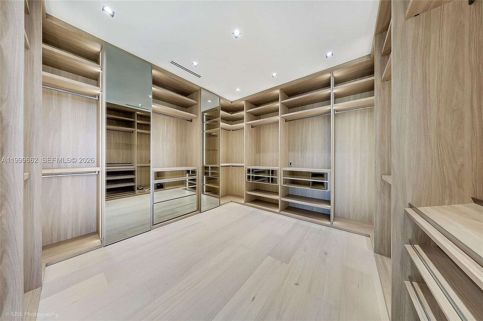 This spacious walk-in closet features custom light wood cabinetry with a mix of open shelving, hanging rods, and integrated drawer units. The room is accented by mirrored cabinet doors that reflect the space, enhancing the sense of depth and brightness. The clean, minimalist design is complemented by recessed ceiling lighting and light-toned wood flooring.