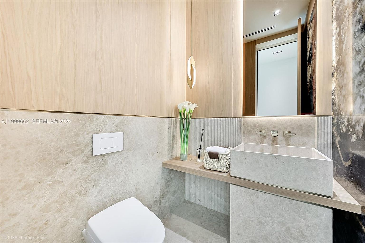 This modern powder room features a minimalist aesthetic with light wood paneling and neutral stone surfaces. A rectangular stone vessel sink sits atop a floating wood vanity, complemented by a wall-mounted faucet and a sleek, frameless mirror. The space is accented by a delicate pendant light and a small basket of towels, creating a sophisticated and serene atmosphere.