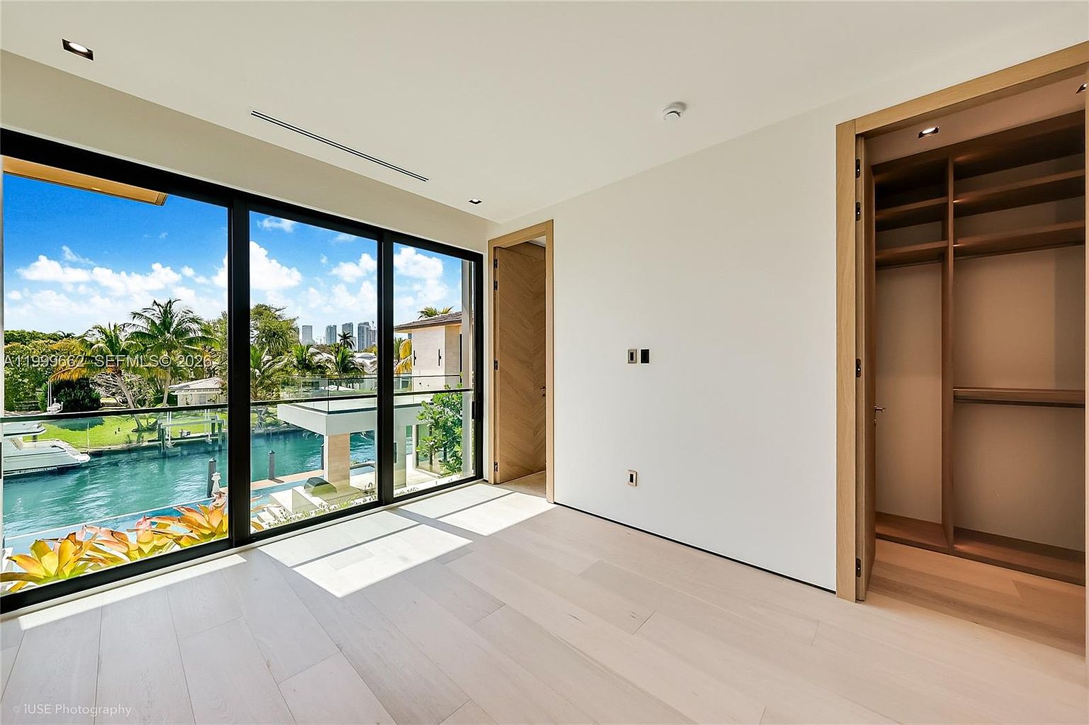 This bright, minimalist bedroom features light wood flooring and crisp white walls, creating an airy and modern atmosphere. A large expanse of floor-to-ceiling glass provides a stunning view of the canal, a private dock, and the surrounding tropical landscape. To the right, an open-concept walk-in closet with built-in shelving offers convenient storage, while a doorway leads to an adjacent room.