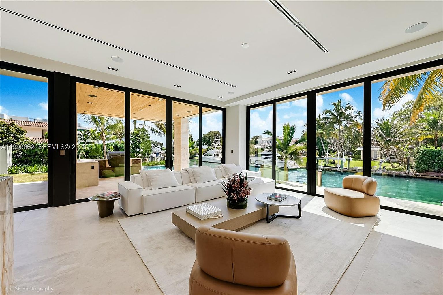 This bright, contemporary living room features a minimalist white sectional sofa and two tan leather armchairs arranged around a low, light-toned wooden coffee table. The space is defined by expansive floor-to-ceiling glass walls that provide an unobstructed view of a private canal, a swimming pool, and lush tropical landscaping. The neutral color palette and clean lines create a serene, airy atmosphere that seamlessly blends indoor and outdoor living.