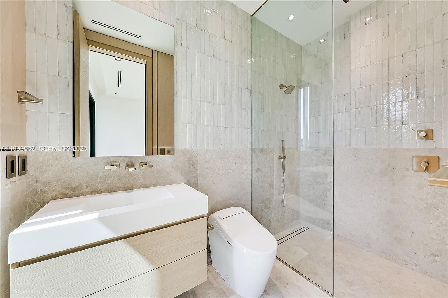 This modern bathroom features a sleek, light wood vanity with a white integrated sink and a large, rectangular mirror. To the right, a glass-enclosed walk-in shower is finished with textured, light-colored wall tiles and stone flooring. The space is illuminated by recessed lighting, creating a clean, minimalist, and high-end aesthetic.