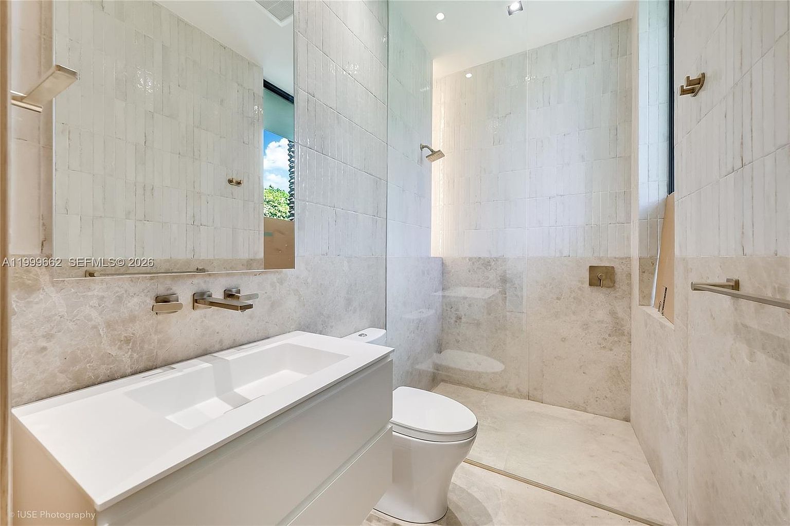 This modern bathroom features a sleek, wall-mounted white vanity with a rectangular sink and a minimalist faucet. The space is finished with light-toned stone wall tiles and a glass-enclosed walk-in shower area. A large, frameless mirror reflects the room, while a white toilet is positioned neatly beside the shower enclosure.