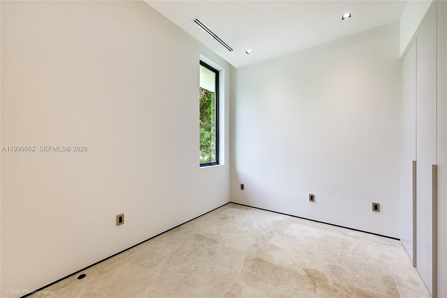 This minimalist, unfurnished room features neutral white walls and light-toned stone flooring. A tall, narrow window provides natural light and a view of greenery outside, while a wall of built-in cabinetry with sleek, handle-less doors offers ample storage. The space is clean and modern, presenting a versatile area suitable for a bedroom or home office.