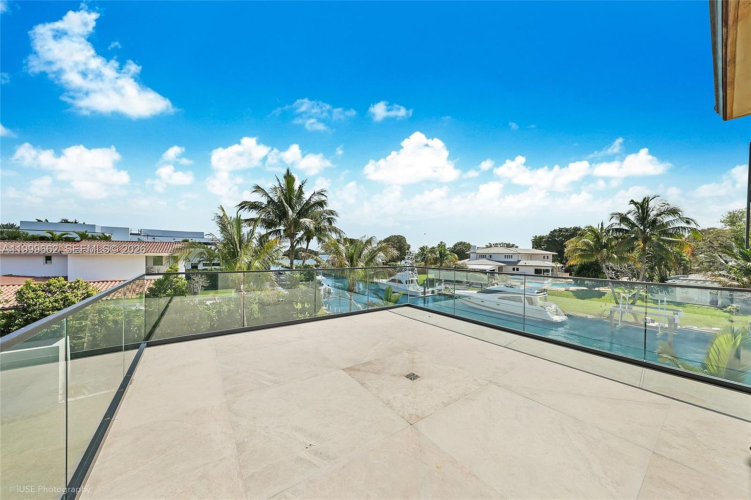 This expansive, open-air balcony features large, light-toned stone tiles and a sleek, frameless glass railing that provides an unobstructed view of the waterfront. The perspective captures a serene canal scene with luxury yachts docked nearby, surrounded by lush tropical landscaping and clear blue skies. It offers a sophisticated, modern outdoor living space perfect for enjoying the coastal scenery.