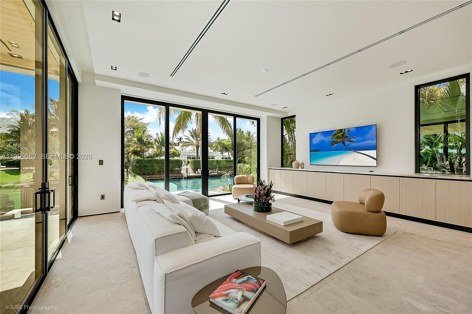 This bright and airy living room features a minimalist design with a large white sofa, a low-profile coffee table, and two tan accent chairs. The space is defined by expansive glass walls that provide a seamless transition to the outdoor pool area and lush tropical landscaping. A large television is mounted above a long, light wood media console, creating a clean and sophisticated entertainment space.