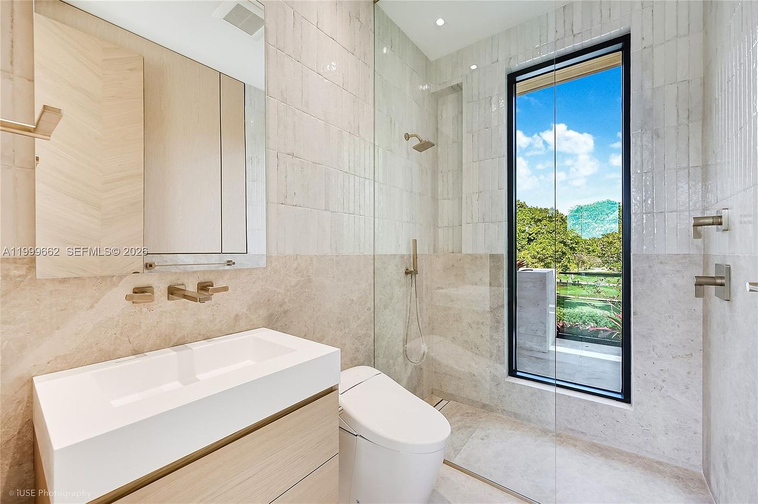 This modern bathroom features a sleek, wall-mounted vanity with a light wood finish and a rectangular vessel sink paired with gold-toned fixtures. A glass-enclosed shower area includes a handheld sprayer and a rainfall showerhead, set against a backdrop of textured light-colored wall tiles and stone flooring. A large window provides a clear view of the lush green outdoors, creating a bright and airy atmosphere.