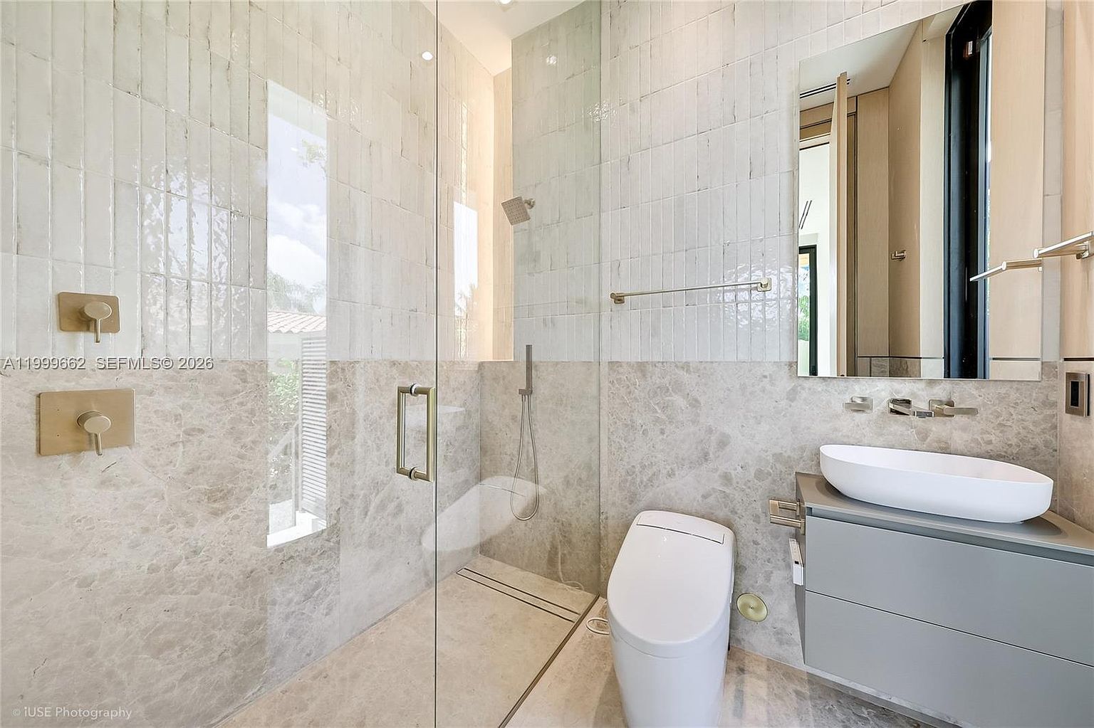 This modern bathroom features a sleek, minimalist design with a glass-enclosed walk-in shower and a wall-mounted toilet. The space is finished with a combination of textured vertical tiles on the upper walls and smooth stone tiles on the lower walls and floor. A floating vanity with a vessel sink and a large, frameless mirror completes the contemporary aesthetic.