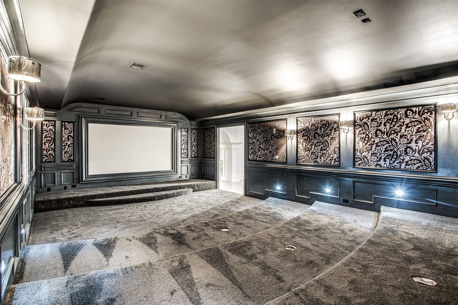 This dedicated home theater room features tiered seating platforms and a large projection screen framed by ornate, dark wood paneling. The walls are adorned with decorative patterned panels and wall sconces, creating a sophisticated and moody atmosphere. The room is carpeted throughout, and the perspective from the back of the theater highlights the spacious layout designed for an immersive cinematic experience.
