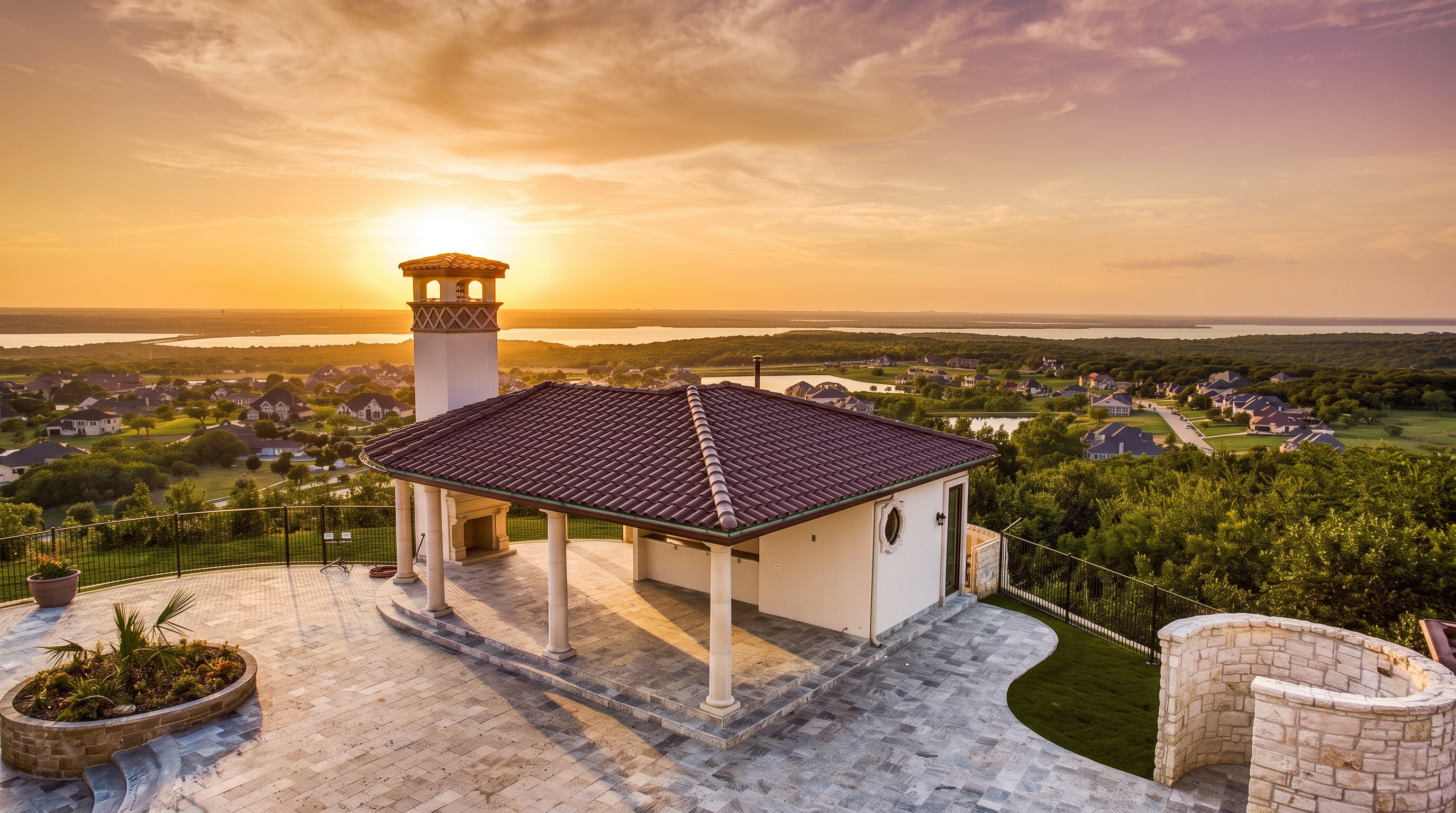 This elevated outdoor living space features a covered pavilion with a prominent stone chimney and a tiled roof, overlooking a sprawling landscape of rolling hills and distant water. The expansive stone-paved patio provides a generous area for entertaining, framed by a decorative stone wall and black metal fencing. The perspective is captured from an elevated vantage point, emphasizing the panoramic views and the luxurious, open-air design of the property.