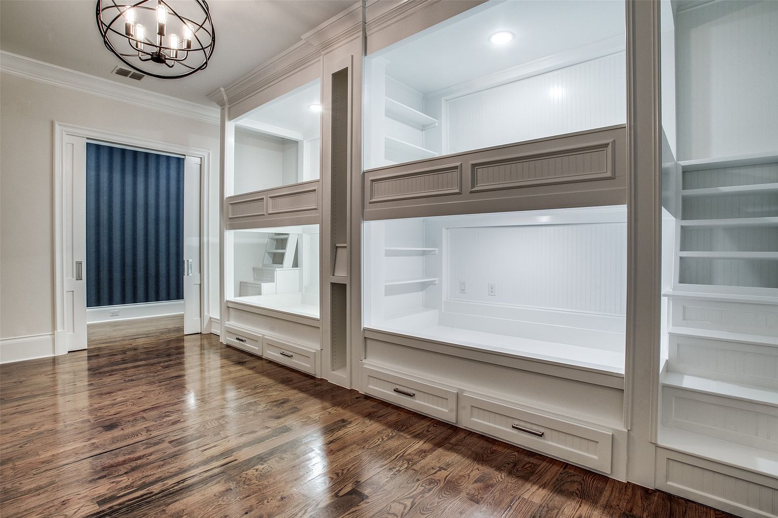 This bedroom features custom-built, white-painted bunk beds with integrated shelving and storage drawers, creating a highly functional and organized sleeping area. The room is finished with polished hardwood flooring and a modern, spherical metal chandelier that adds a contemporary touch. A sliding door leads to an adjacent space, showcasing a clean, bright aesthetic suitable for a children's room or guest suite.